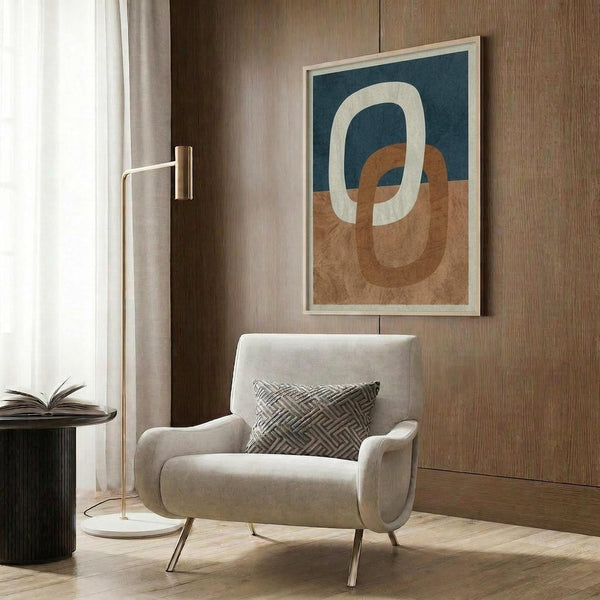 May include: Abstract art print with interlocking oval rings in navy blue, terracotta orange, and cream colors, displayed in light wood frame on wood paneled wall above beige upholstered armchair with geometric patterned pillow, brass floor lamp, and dark side table with open book in modern living room setting