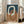 May include: Abstract geometric poster with interlocking oval rings in cream, terracotta orange, and navy blue colors, displayed in light wood frame leaning against white wall next to beige upholstered chair in bright living room with large windows and natural sunlight streaming across hardwood floors.
