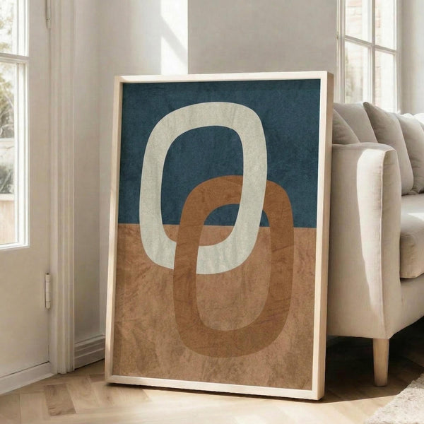 May include: Abstract geometric poster with interlocking oval rings in cream, terracotta orange, and navy blue colors, displayed in light wood frame leaning against white wall next to beige upholstered chair in bright living room with large windows and natural sunlight streaming across hardwood floors.
