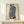 May include: Framed abstract fashion illustration displayed on light wooden shelf against cream wall. Artwork shows stylized figure in blue pinstriped top and leopard print pants, holding brown purse. Figure has dark hair and warm skin tone against soft gradient background. Natural wood frame complements minimalist aesthetic. Small white wrapped gift with evergreen sprig sits on shelf beside frame.