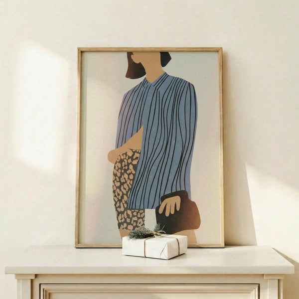 May include: Framed abstract fashion illustration displayed on light wooden shelf against cream wall. Artwork shows stylized figure in blue pinstriped top and leopard print pants, holding brown purse. Figure has dark hair and warm skin tone against soft gradient background. Natural wood frame complements minimalist aesthetic. Small white wrapped gift with evergreen sprig sits on shelf beside frame.