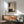 May include: Abstract art print in wooden frame hanging above mid-century modern credenza with table lamp. Artwork features black brushstrokes, pink and beige geometric shapes, and vertical dots on cream background. Green upholstered chair with wooden arms sits nearby. Bright natural light creates shadows on white wall in contemporary living room setting.