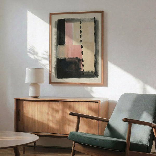 May include: Abstract art print in wooden frame hanging above mid-century modern credenza with table lamp. Artwork features black brushstrokes, pink and beige geometric shapes, and vertical dots on cream background. Green upholstered chair with wooden arms sits nearby. Bright natural light creates shadows on white wall in contemporary living room setting.