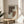 May include: Abstract geometric art print in wooden frame on round wooden table next to beige upholstered chair. Artwork features earthy brown and tan shapes with dotted stippled texture pattern on cream background. Soft natural lighting creates window shadows across neutral-toned room setting with plant leaves visible in corner.