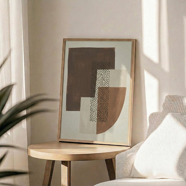 May include: Abstract geometric art print in wooden frame on round wooden table next to beige upholstered chair. Artwork features earthy brown and tan shapes with dotted stippled texture pattern on cream background. Soft natural lighting creates window shadows across neutral-toned room setting with plant leaves visible in corner.