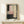 May include: Abstract artwork in natural wood frame displayed on light wooden surface with small wrapped gift. The painting features geometric blocks in soft pink, cream, and charcoal gray with vertical row of black dots down center, set against minimalist white wall background.