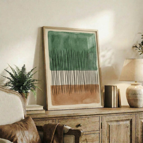 May include: Abstract watercolor artwork in light wood frame displaying green wash at top, vertical black line pattern in middle, and brown wash at bottom. Framed print sits on rustic wooden sideboard next to fern plant, ceramic vase, and stacked books in neutral-toned living room setting.