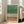 May include: Framed abstract artwork with green watercolor wash at top, vertical black lines creating grass-like texture, and warm beige wash at bottom. Light wood frame displayed against white wall next to beige upholstered sofa in bright living room with large windows and natural light.