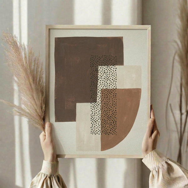 May include: Person in cream sweater holding framed abstract geometric art print featuring overlapping shapes in brown, terracotta, and cream with black polka dot patterns, displayed alongside dried wheat stems against white curtained window background