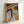 Framed artwork leaning against white wall showing a figure with long dark hair wearing blue and white striped clothing, painted in warm earth tones with stylized wavy lines above. The piece features a brown background with geometric patterns and is displayed in a light wood frame.