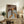 May include: Framed portrait artwork depicting a figure in blue and white striped garment holding an orange cat, with wavy white lines against dark brown background, displayed on rustic wooden sideboard alongside books, ceramic vase, and fern plant in bright, modern living room setting with cream armchair visible.