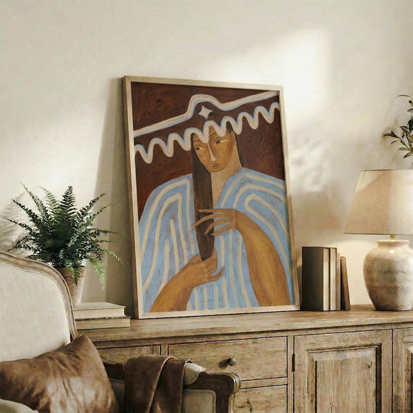 May include: Framed portrait artwork depicting a figure in blue and white striped garment holding an orange cat, with wavy white lines against dark brown background, displayed on rustic wooden sideboard alongside books, ceramic vase, and fern plant in bright, modern living room setting with cream armchair visible.