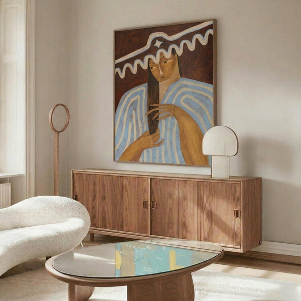 May include: Framed artwork showing a woman with long dark hair in a blue and white striped garment holding what appears to be a mirror, painted in warm earth tones with decorative white curved elements in the background. The print is displayed above a wooden sideboard with brass handles in a modern living room setting with white upholstered chair, oval glass coffee table, and contemporary lighting fixtures.