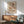 May include: Abstract geometric art print with overlapping circles and curved shapes in warm earth tones of brown, orange, peach and mint green, displayed in wooden frame above mid-century modern credenza. Scene shows sage green upholstered armchair with wooden arms, white ceramic table lamp, and natural sunlight casting shadows on white wall.