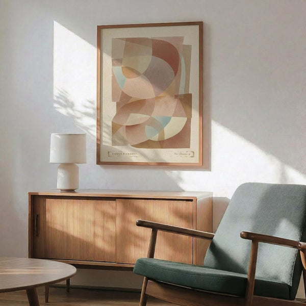 May include: Abstract geometric art print with overlapping circles and curved shapes in warm earth tones of brown, orange, peach and mint green, displayed in wooden frame above mid-century modern credenza. Scene shows sage green upholstered armchair with wooden arms, white ceramic table lamp, and natural sunlight casting shadows on white wall.