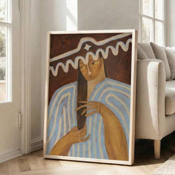 May include: A framed art print leaning against a beige armchair in a bright living room with white walls and windows. The artwork depicts two figures with golden-brown skin tones wearing blue and white striped garments, set against a dark brown background with flowing white design elements above. The print is displayed in a light wood frame on hardwood flooring.