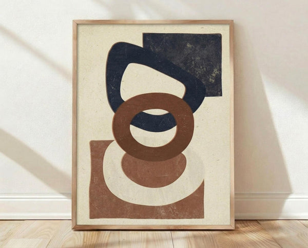 May include: Framed abstract geometric art print with overlapping shapes in navy blue, terracotta brown, and cream colors on textured beige background. Wooden frame sits on light wood flooring against white wall with natural lighting creating soft shadows.