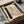 May include: Framed abstract art print in black frame sitting in cardboard shipping box with protective tissue paper. Artwork features pink and beige rectangular color blocks, black brushstroke, and row of small black dots connected by curved line. Wooden dowels visible alongside packaging materials.