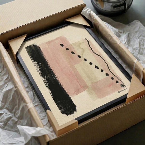 May include: Framed abstract art print in black frame sitting in cardboard shipping box with protective tissue paper. Artwork features pink and beige rectangular color blocks, black brushstroke, and row of small black dots connected by curved line. Wooden dowels visible alongside packaging materials.