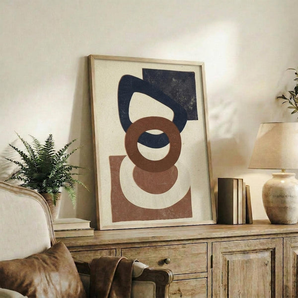 May include: Abstract geometric art print in light wood frame leaning against white wall on rustic wooden sideboard. Artwork features navy blue and terracotta brown organic shapes on cream background. Scene styled with fern plant, ceramic vase, stacked books, and brown leather chair corner visible.