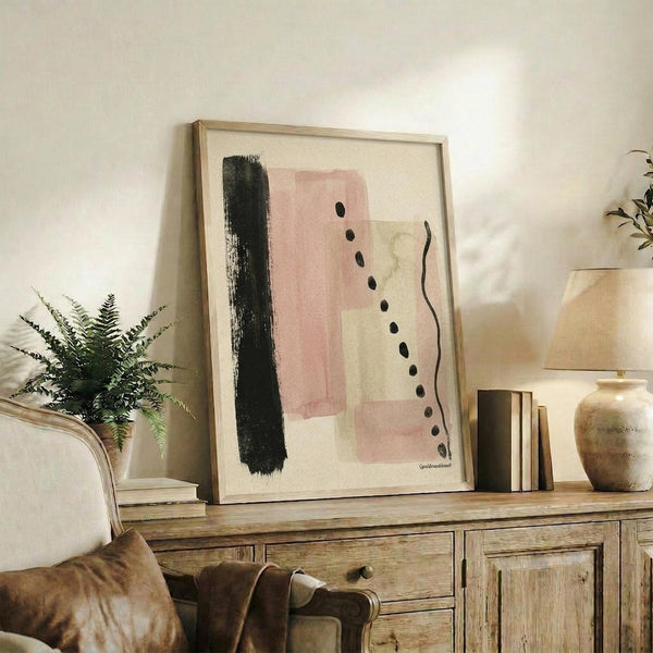 May include: Abstract art print in light wood frame displayed on rustic wooden console table beside fern plant and books. Artwork features soft pink watercolor blocks, black brushstroke, dotted line, and wavy line on cream background. Cozy living room setting with beige chair, throw blanket, and ceramic vases creating warm, modern home decor styling.