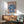 May include: Framed artwork showing a woman with flowing blue hair or fabric billowing around her against a pink background, displayed above a wooden sideboard with table lamp in a modern living room with mint green armchair and natural lighting creating shadow patterns on white wall.