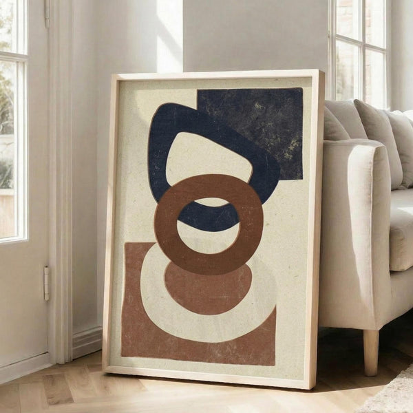 May include: Large framed abstract geometric art print leaning against white wall next to beige armchair in bright living room. Artwork features overlapping organic shapes in navy blue, terracotta brown, and cream on textured off-white background within light wood frame. Natural sunlight streams through tall windows creating soft shadows on hardwood floors.
