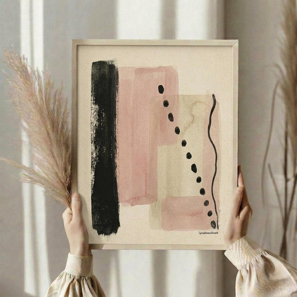 May include: Person holding framed abstract art print featuring vertical black brushstroke, pink and beige geometric shapes, and diagonal line of black dots on cream background. Light wood frame, held alongside dried pampas grass stems against soft beige curtains in natural lighting.