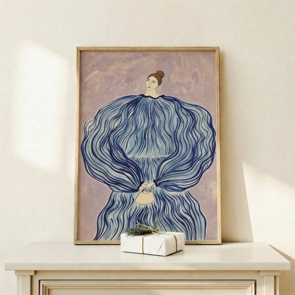 May include: Framed artwork featuring a woman with dark hair in a bun wearing a voluminous blue flowing dress or cape with wavy striped patterns, set against a soft purple background. The piece is displayed in a light wood frame on a cream-colored shelf next to a small white wrapped gift box with greenery on top.
