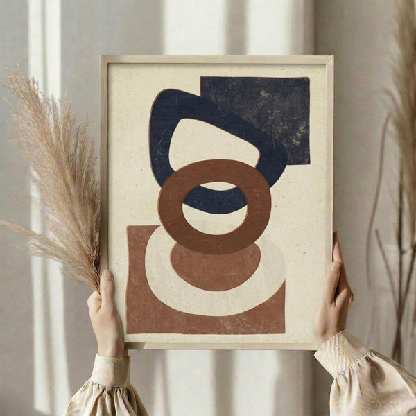 May include: Person holding framed abstract geometric art print featuring overlapping shapes in navy blue, terracotta brown, and cream colors on beige background, displayed in light wood frame against neutral curtained window with dried pampas grass stems visible to the left