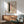 May include: Abstract art print with pink rectangles, dark green brushstroke, black dots, and curved line on cream background, displayed in wood frame above mid-century modern credenza. Sage green upholstered chair with wooden arms and small round table visible. Warm natural lighting creates shadows on white wall.
