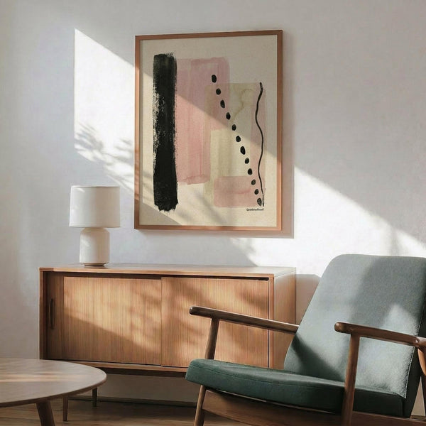 May include: Abstract art print with pink rectangles, dark green brushstroke, black dots, and curved line on cream background, displayed in wood frame above mid-century modern credenza. Sage green upholstered chair with wooden arms and small round table visible. Warm natural lighting creates shadows on white wall.