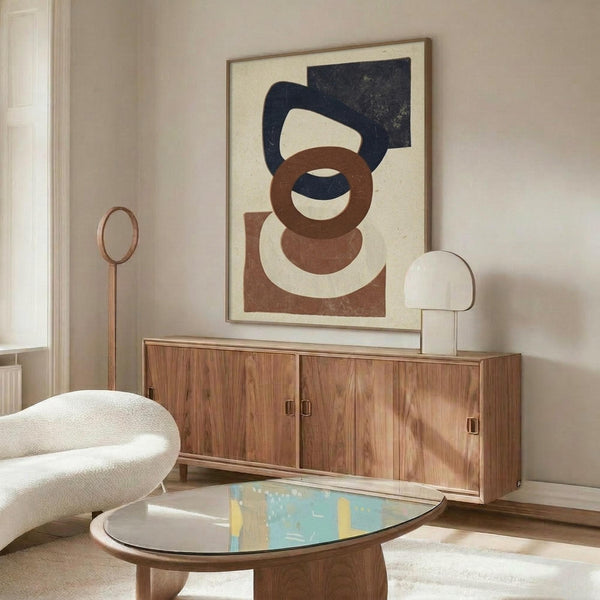 May include: Abstract geometric art print in cream, navy blue, and brown tones featuring overlapping circles and curved shapes, displayed in thin wooden frame above walnut credenza in modern living room with white curved chair, brass floor lamp, glass coffee table, and natural lighting creating warm shadows on neutral walls.