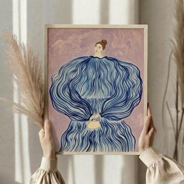 May include: Hands holding framed art print showing woman with flowing blue dress or hair against pink background, displayed with dried pampas grass stems in soft natural lighting with white curtains visible in background