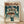 May include: Framed anime poster displayed on wooden floor against white wall. Shows character in blue bodysuit holding umbrella in rain with large mecha robot behind them. Japanese text at top in orange letters. Artwork has teal and orange color scheme with dramatic lighting effects. Natural wood frame with visible grain pattern.
