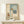 May include: Framed art print displayed on light wood console showing minimalist illustration of figure in white dress and wide-brimmed hat standing before arched mirror reflecting ocean view, with wooden chair and potted plant nearby. Soft neutral color palette with sage green and cream tones. Small wrapped gift with pine sprig sits on console surface.