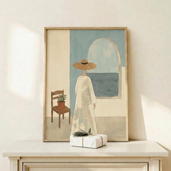 May include: Framed art print displayed on light wood console showing minimalist illustration of figure in white dress and wide-brimmed hat standing before arched mirror reflecting ocean view, with wooden chair and potted plant nearby. Soft neutral color palette with sage green and cream tones. Small wrapped gift with pine sprig sits on console surface.