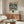 May include: Framed anime poster featuring a young character with umbrella standing before a large mecha robot in rain, displayed above a mid-century modern walnut credenza in a minimalist living room with white walls, cream chair, glass coffee table, and warm natural lighting.