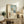 May include: Framed poster print leaning against white wall on rustic wooden sideboard, featuring minimalist artwork of figure in white dress and wide-brimmed hat looking toward blue window with small chair and plant. Warm neutral room setting with fern plant, ceramic vase, books, and beige upholstered furniture visible.