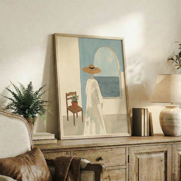 May include: Framed poster print leaning against white wall on rustic wooden sideboard, featuring minimalist artwork of figure in white dress and wide-brimmed hat looking toward blue window with small chair and plant. Warm neutral room setting with fern plant, ceramic vase, books, and beige upholstered furniture visible.