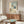 May include: Modern minimalist artwork in thin wooden frame hanging above walnut credenza, showing woman in wide-brimmed hat and white dress standing before arched window overlooking blue sea, with small wooden chair and potted plant, displayed in bright living room with white curved chair and oval glass coffee table