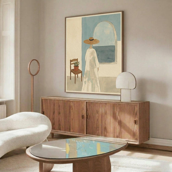 May include: Modern minimalist artwork in thin wooden frame hanging above walnut credenza, showing woman in wide-brimmed hat and white dress standing before arched window overlooking blue sea, with small wooden chair and potted plant, displayed in bright living room with white curved chair and oval glass coffee table