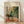 May include: Framed Henri Matisse exhibition poster in light wood frame leaning against white wall on wooden floor. Colorful Fauve-style painting shows teacup and saucer on pink table with lush green tropical plants and window view. Vibrant brushstrokes in yellows, greens, oranges. Gallery exhibition text visible at top right. Soft natural lighting creates gentle shadows on white textured wall.