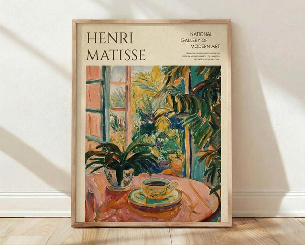 May include: Framed Henri Matisse exhibition poster in light wood frame leaning against white wall on wooden floor. Colorful Fauve-style painting shows teacup and saucer on pink table with lush green tropical plants and window view. Vibrant brushstrokes in yellows, greens, oranges. Gallery exhibition text visible at top right. Soft natural lighting creates gentle shadows on white textured wall.