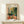 May include: Framed Henri Matisse exhibition poster featuring vibrant tropical interior scene with potted plants, teacup on round table, and window view. Colorful brushstrokes in greens, oranges, and yellows. Natural wood frame on light wooden shelf beside small wrapped gift with evergreen sprig against cream wall.