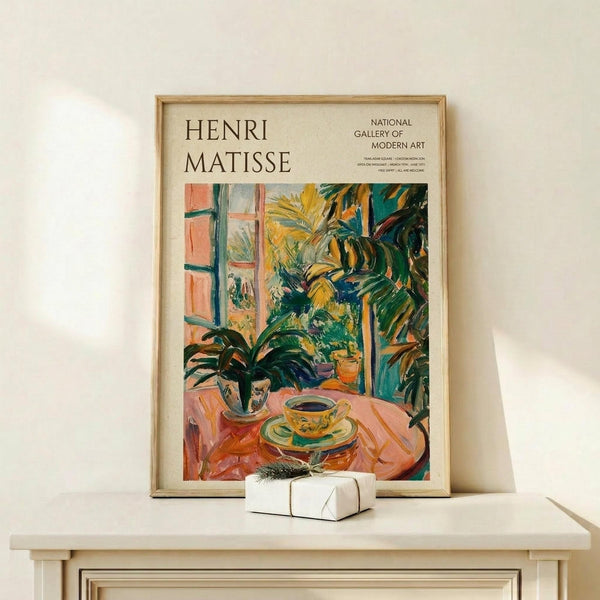 May include: Framed Henri Matisse exhibition poster featuring vibrant tropical interior scene with potted plants, teacup on round table, and window view. Colorful brushstrokes in greens, oranges, and yellows. Natural wood frame on light wooden shelf beside small wrapped gift with evergreen sprig against cream wall.