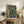 May include: Framed poster featuring an anime-style illustration of a pilot character standing in front of a large green mech robot in a futuristic cityscape with Japanese text at the top, displayed on a rustic wooden sideboard next to a fern plant, ceramic vase, and books in a cozy living room setting with beige chair visible