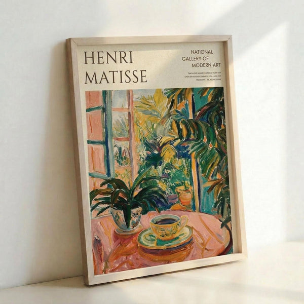 May include: Framed Henri Matisse exhibition poster leaning against white wall. Colorful artwork shows interior scene with pink round table, teacup and saucer, potted plants, and window view. Vibrant greens, yellows, oranges and pinks in Matisse's signature Fauvist style. Light wooden frame on light wood surface with soft natural lighting creating gentle shadows.