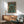 May include: Framed anime-style poster featuring a pilot character standing before a large green mecha robot in a futuristic cityscape setting with Japanese text, displayed above a wooden sideboard with white ceramic lamp in a mid-century modern living room with teal upholstered chair and round coffee table, natural sunlight creating shadows on white walls.