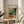 May include: Framed Henri Matisse museum exhibition poster featuring vibrant tropical interior scene with palm plants, teacup on round table, and bright greens, yellows, and oranges. Natural wood frame sits on light wooden side table next to beige upholstered chair with patterned throw pillows. Soft window light creates shadows on neutral wall.